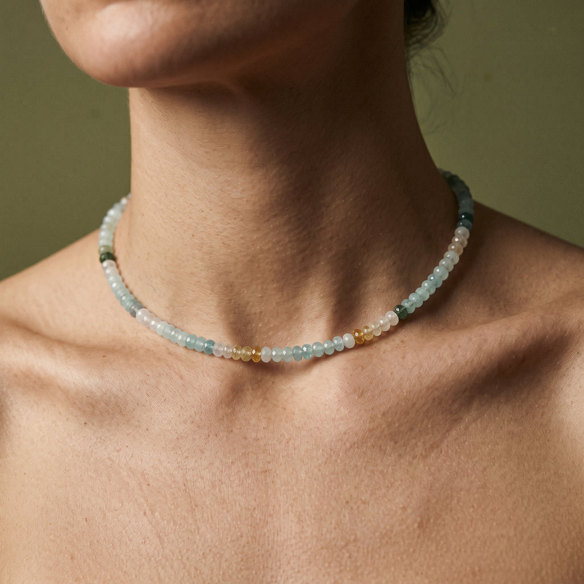 A close-up of a person's neck and upper chest wearing a delicate beaded choker necklace. The necklace features small translucent beads in pale blue, white, and a few warm gold and green accents, set against a soft olive green background.