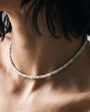 A close-up lifestyle shot of a person's neck and upper chest wearing a thin beaded choker necklace. The beads are a mix of pale sage green, cream, and light blue.