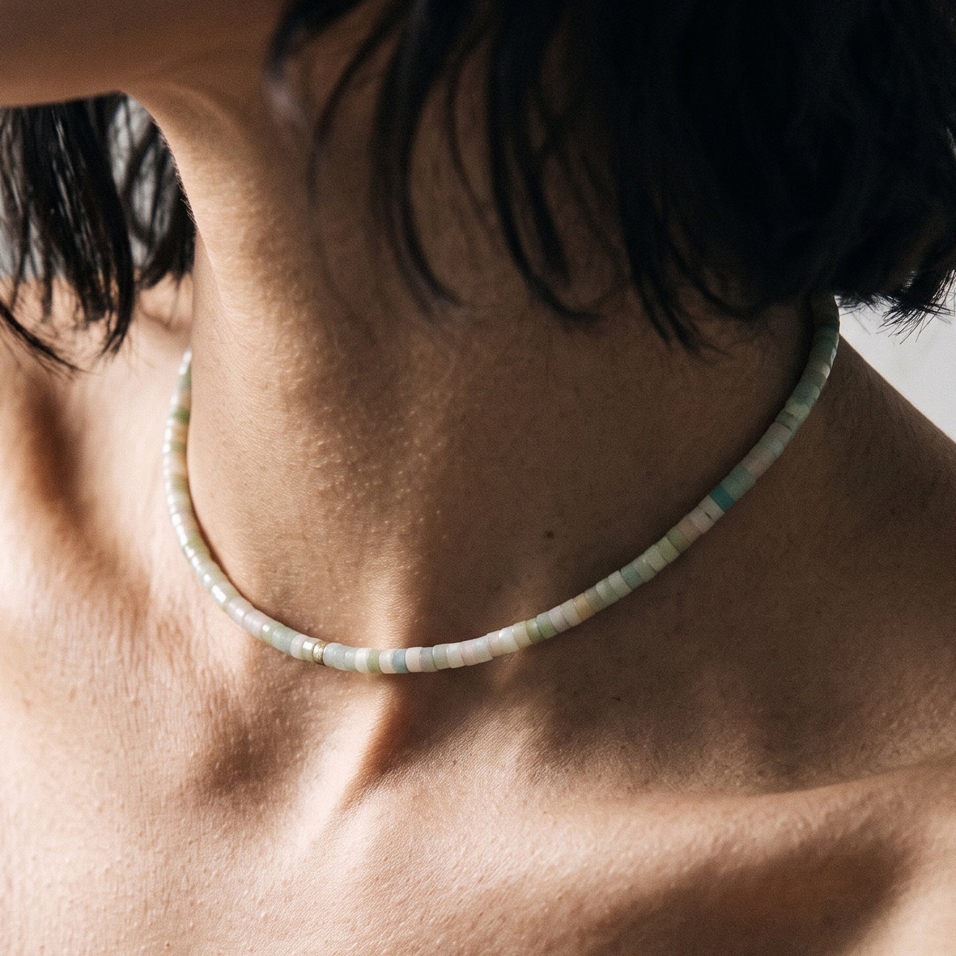 A close-up lifestyle shot of a person's neck and upper chest wearing a thin beaded choker necklace. The beads are a mix of pale sage green, cream, and light blue.