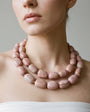 A close-up of a model wearing a double-strand chunky beaded necklace made of pinkish stone beads with natural marbled patterns, paired with a white strapless top against a neutral gray background.