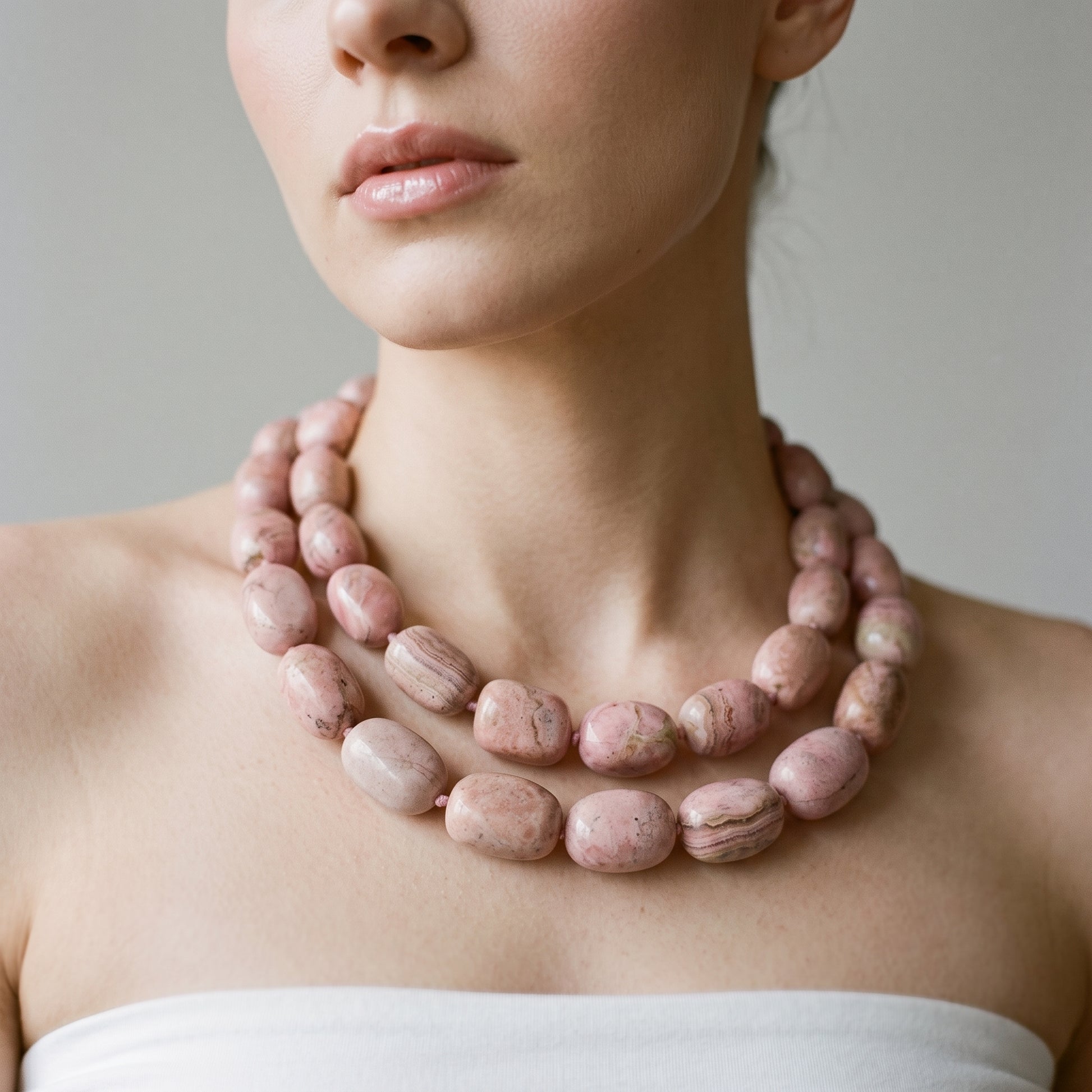 A close-up of a model wearing a double-strand chunky beaded necklace made of pinkish stone beads with natural marbled patterns, paired with a white strapless top against a neutral gray background.