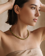 A woman wearing a beaded necklace made of olive green beads, styled with a beige strapless top against a soft beige background.