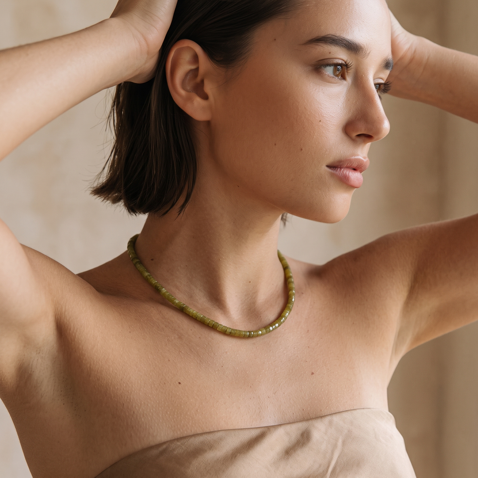 A woman wearing a beaded necklace made of olive green beads, styled with a beige strapless top against a soft beige background.