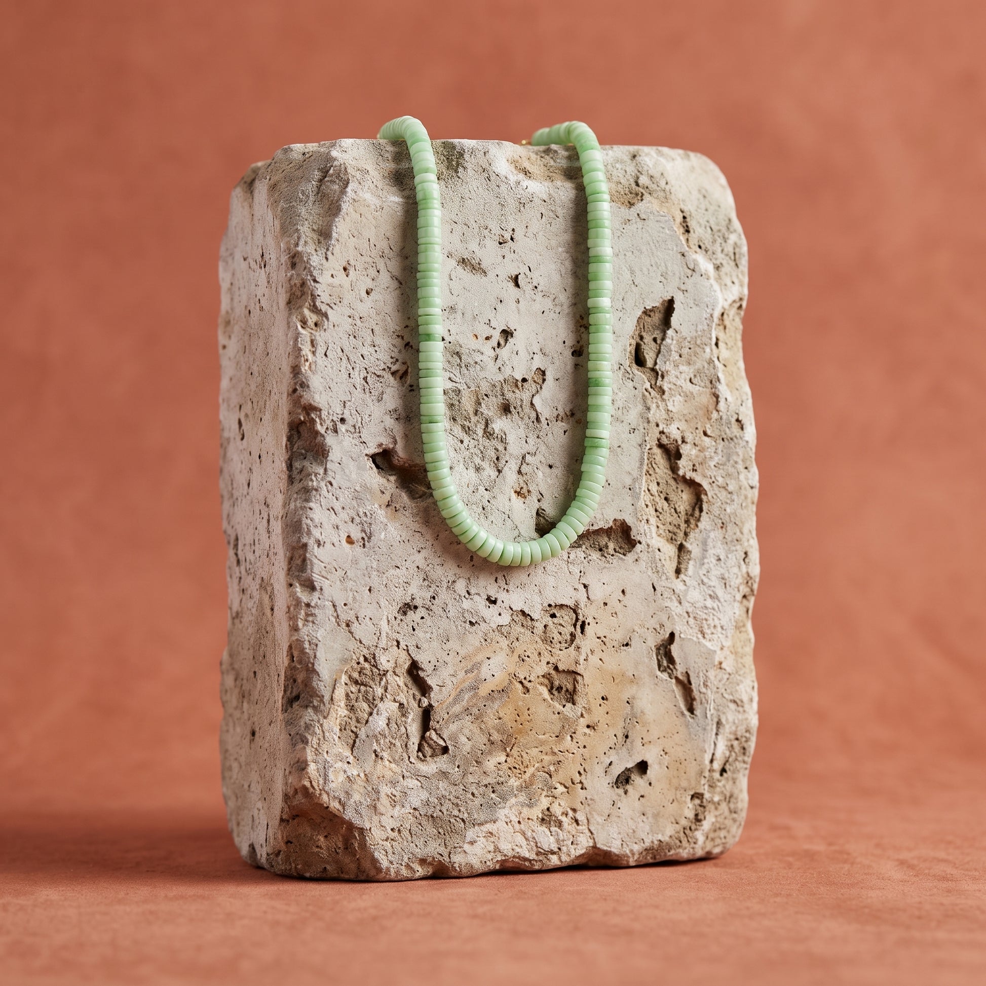 A light green beaded necklace draped over a rough, textured stone block against a warm terracotta background.