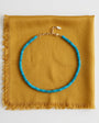 A turquoise beaded necklace with a gold clasp, displayed on a folded mustard yellow fabric with fringe, shot against a clean white background.