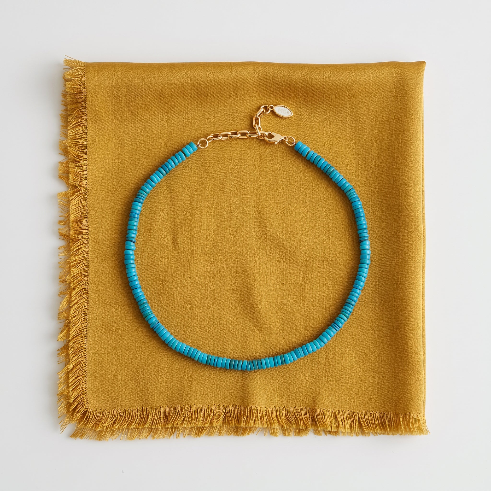A turquoise beaded necklace with a gold clasp, displayed on a folded mustard yellow fabric with fringe, shot against a clean white background.