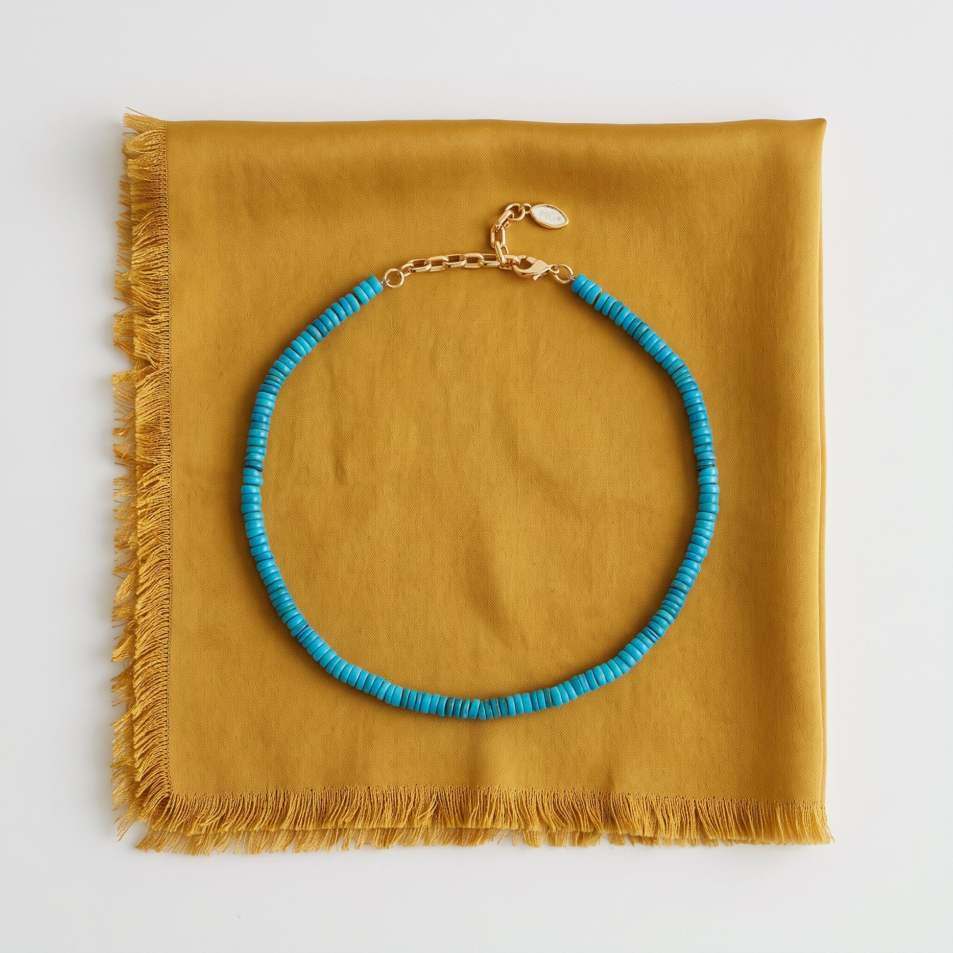 A turquoise beaded necklace with a gold clasp, displayed on a folded mustard yellow fabric with fringe, shot against a clean white background.