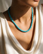 A close-up of a model wearing a turquoise beaded necklace with a chunky, textured design, paired with a cream-colored draped top.