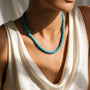 A close-up of a model wearing a turquoise beaded necklace with a chunky, textured design, paired with a cream-colored draped top.