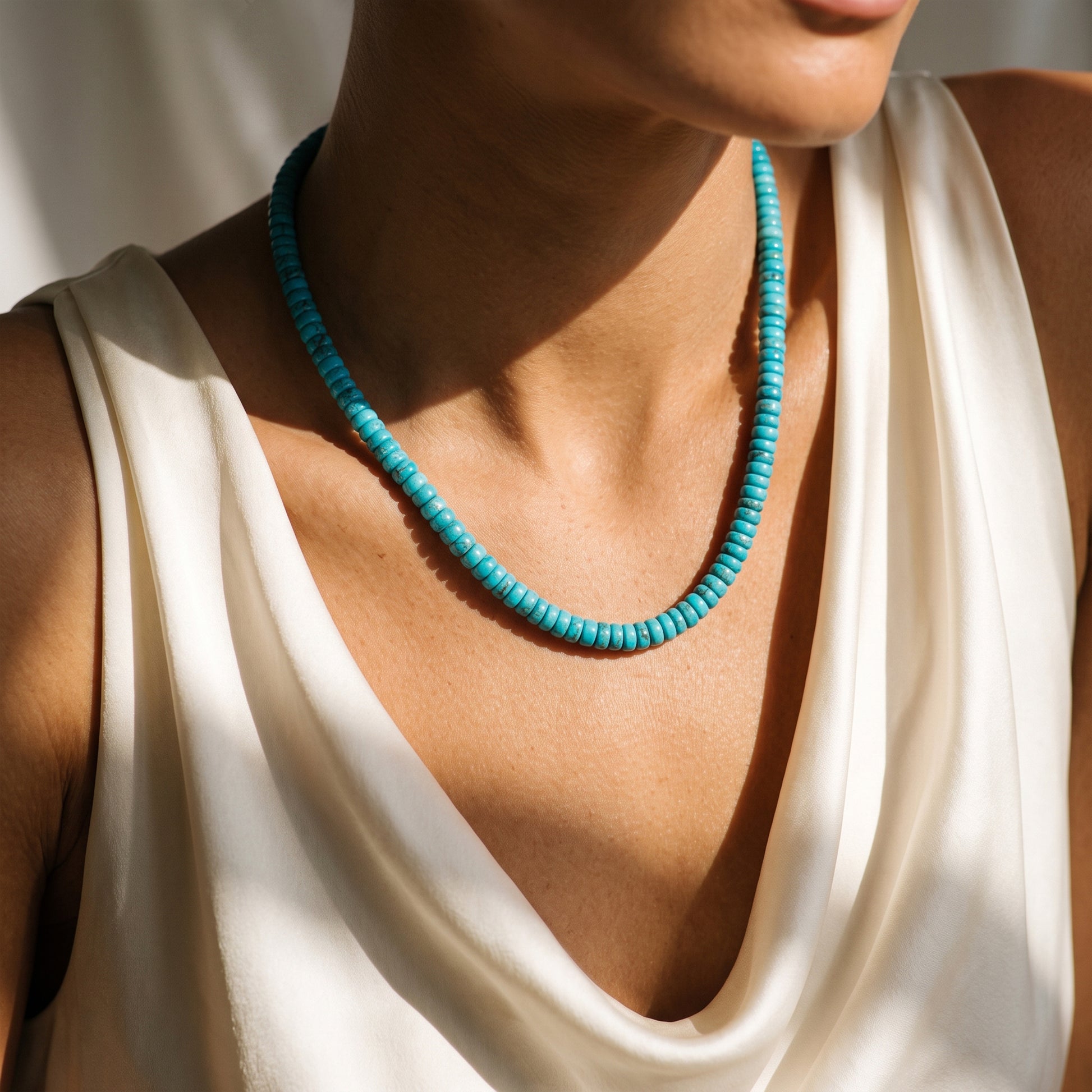 A close-up of a model wearing a turquoise beaded necklace with a chunky, textured design, paired with a cream-colored draped top.