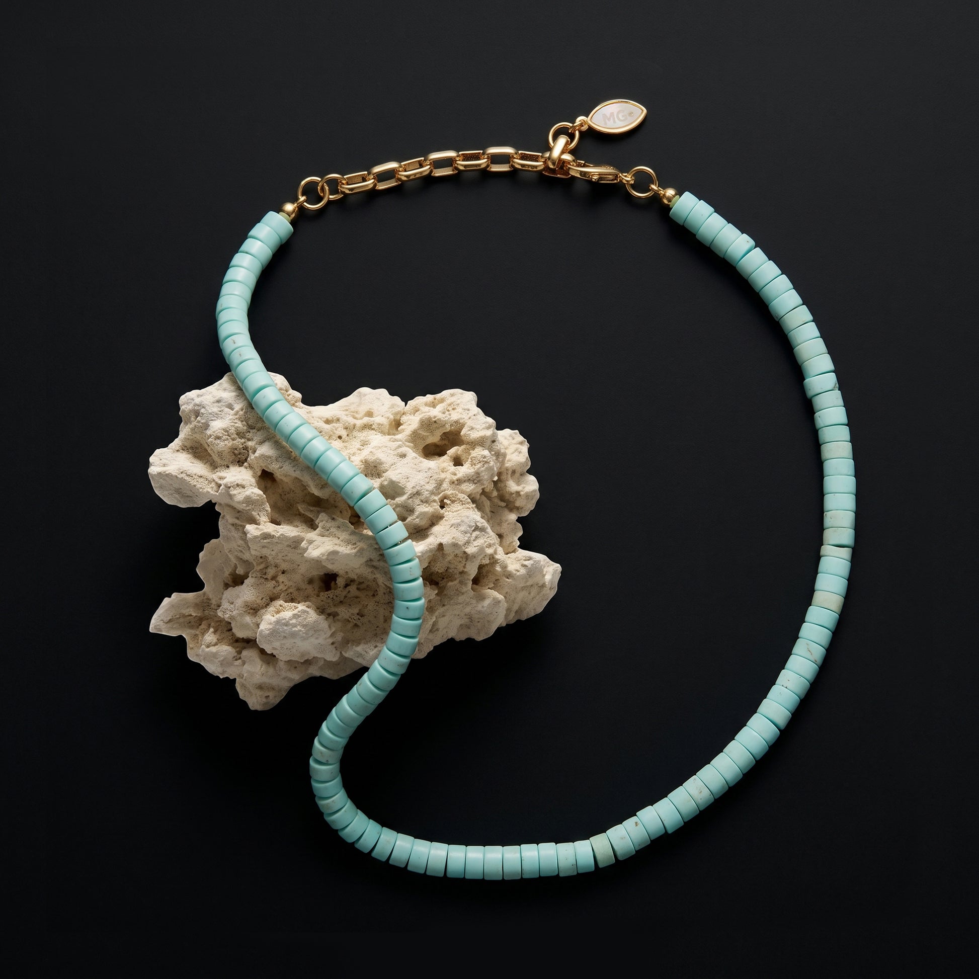 A turquoise beaded necklace with a gold chain clasp, displayed on a textured white rock against a solid black background.