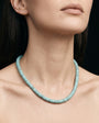 A close-up of a model wearing a light blue-green beaded necklace against a dark background.
