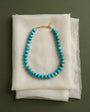 A turquoise beaded necklace with a gold clasp, displayed on a stack of white textured fabric sheets against a dark olive green background.
