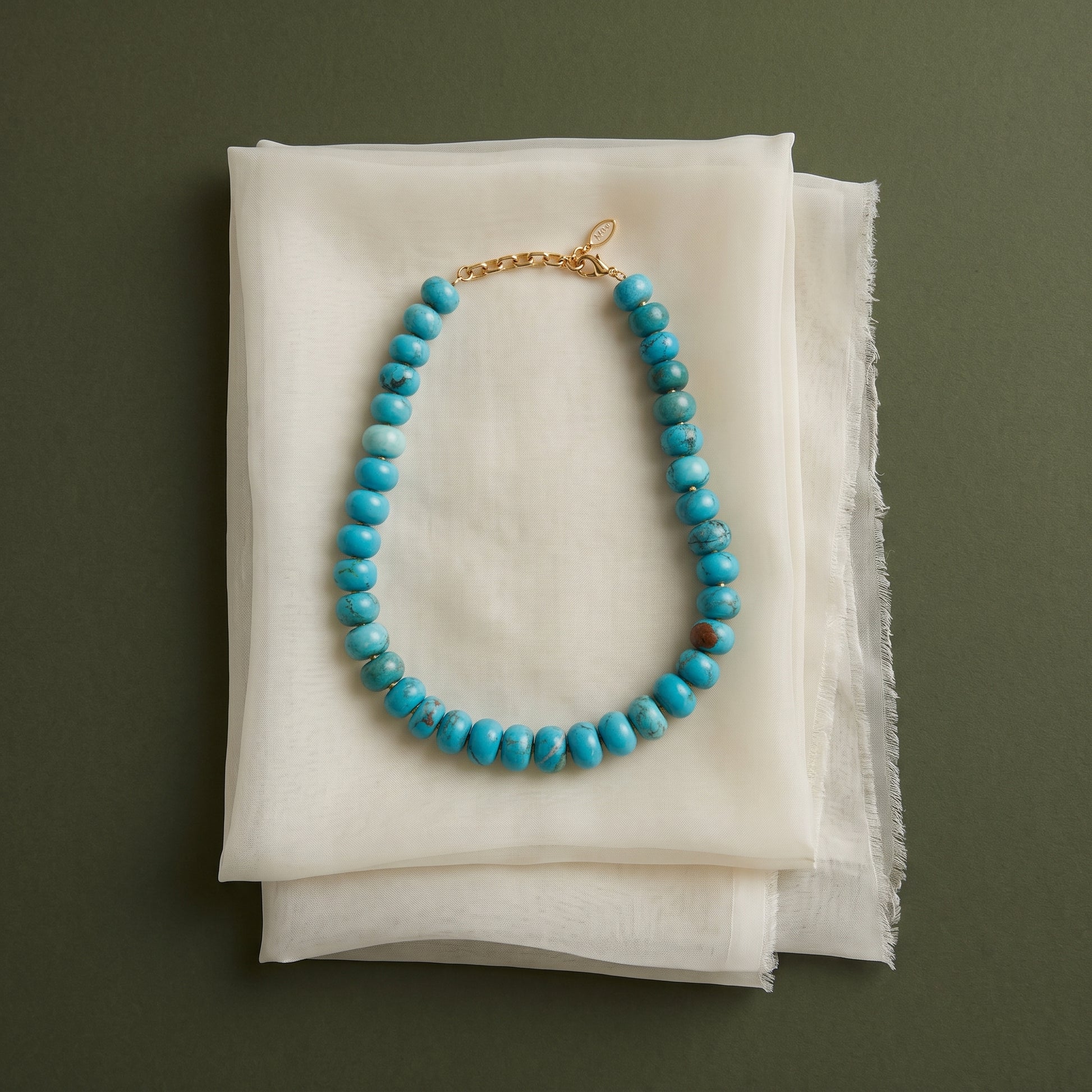 A turquoise beaded necklace with a gold clasp, displayed on a stack of white textured fabric sheets against a dark olive green background.