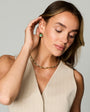 A woman wearing a tennis necklace and matching gold hoop earrings with green gemstones, posing against a solid light gray background.