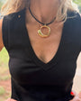A close-up of a woman wearing a black sleeveless V-neck top and a gold-toned double-ring pendant necklace on a black cord. She is also wearing stacked gold bracelets on one hand, with a blurred outdoor background.