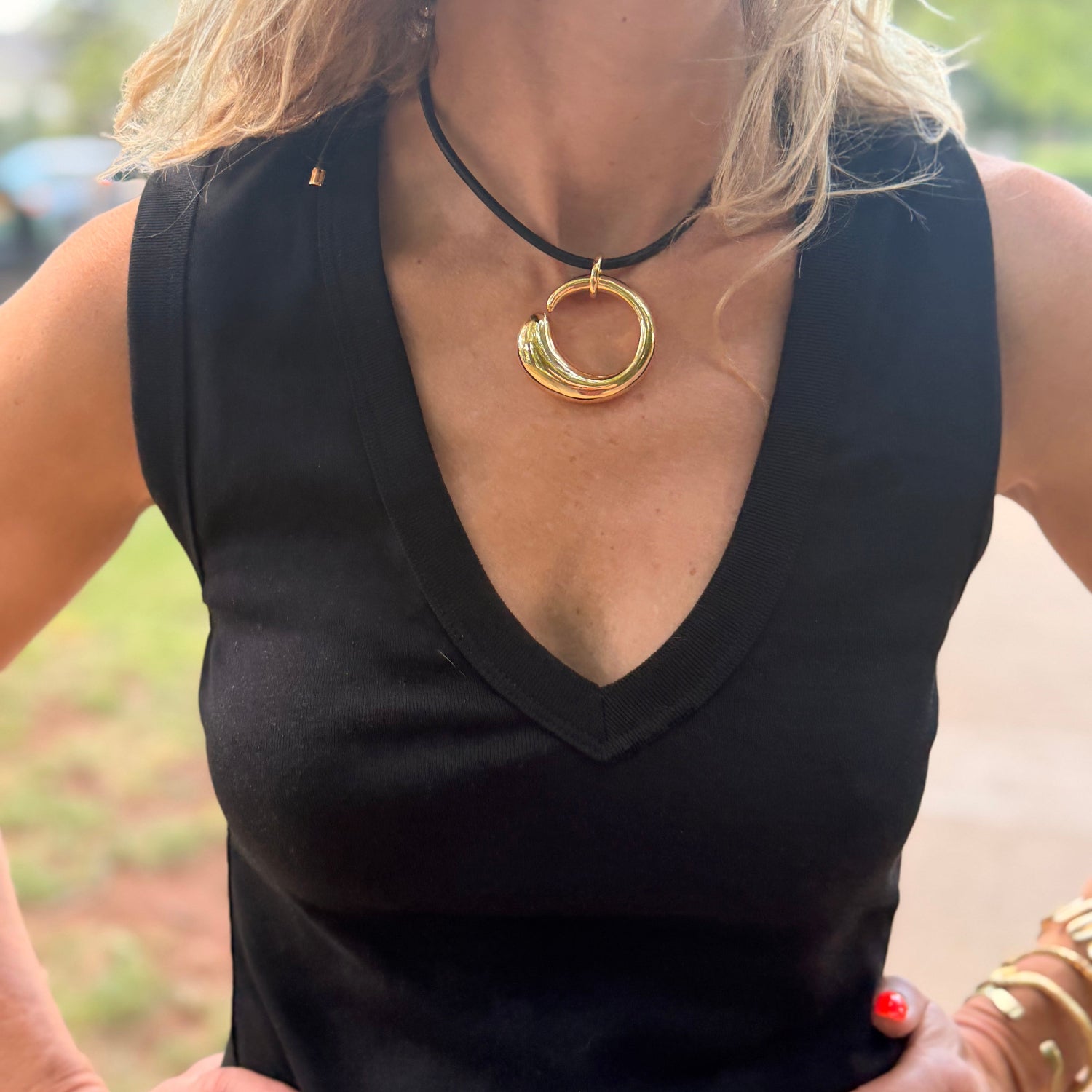 A close-up of a woman wearing a black sleeveless V-neck top and a gold-toned double-ring pendant necklace on a black cord. She is also wearing stacked gold bracelets on one hand, with a blurred outdoor background.