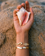 Hand holding a shell on a sandy background with gold bracelets on the wrist.