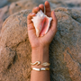 Hand holding a shell on a sandy background with gold bracelets on the wrist.
