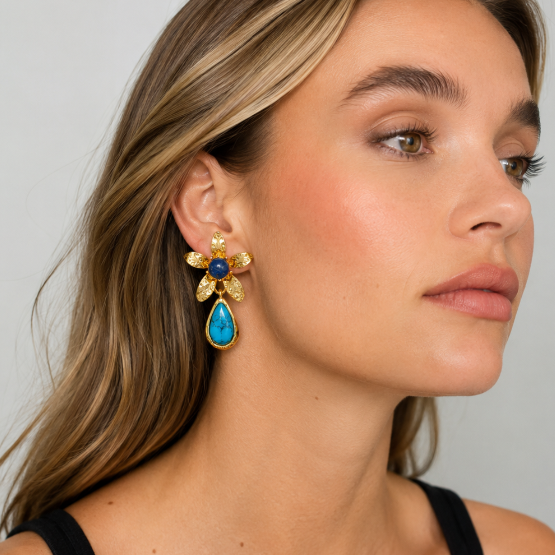 A close-up of a woman wearing a gold flower-shaped dangle earring with a blue teardrop stone and a small dark blue center, against a light gray background.