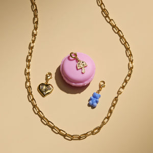 Crystal and Gold Heart Charm, Blue Gummy Bear Charm, and Enamel and Crystal Mushroom Cham Staged with Gold Chain Necklace and Pink Macaroon Cookie on Cream Surface