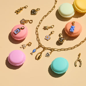 Various Charms and Gold Chain Necklace Staged with Pastel Macaroon Cookies on Flat Cream Surface