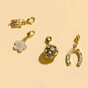 Crystal and Gold Turtle Charm, Flower Charm, Gold Ball with Black and White Evil Eye Charm, and Gold and Crystal Horseshoe Charm on Flat Cream Background