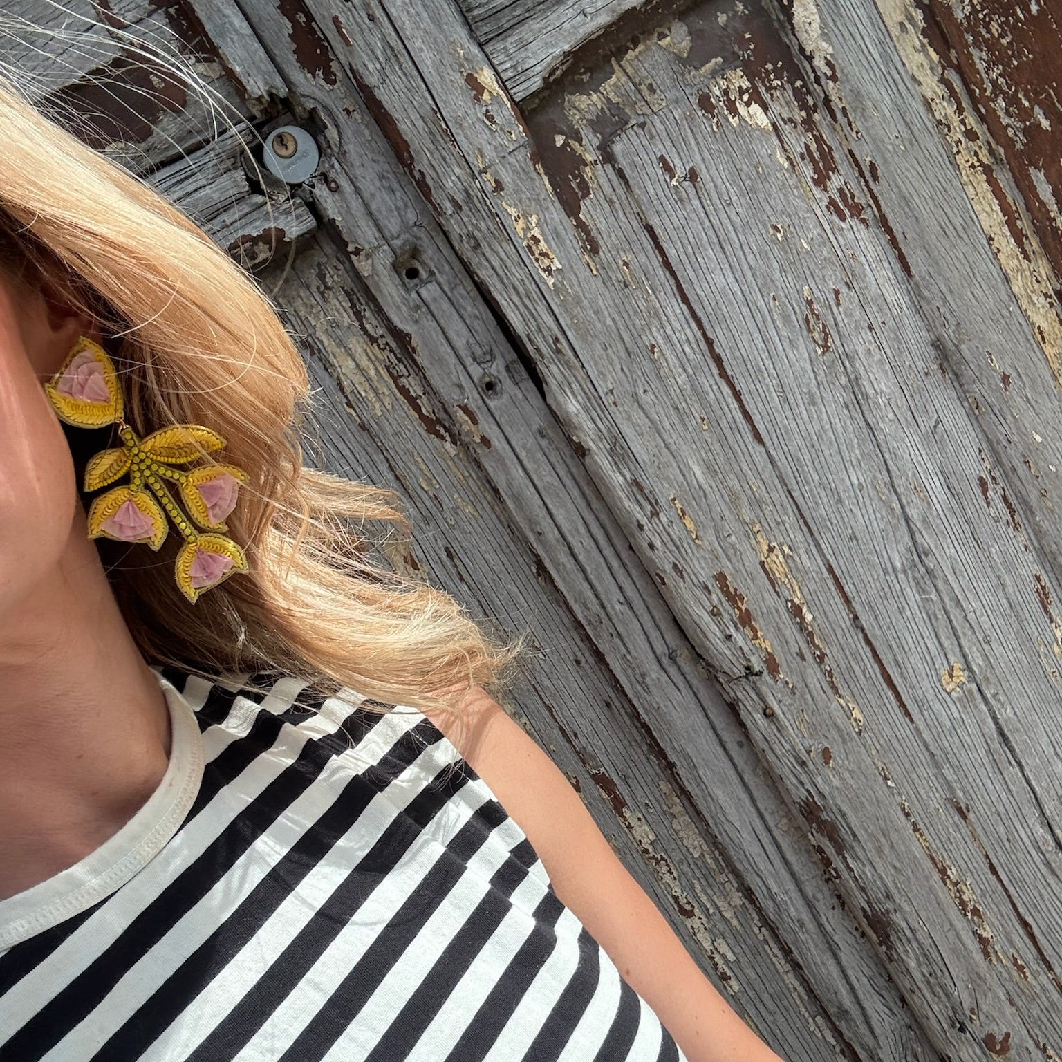 Person wearing a black and white striped shirt with a floral earring, standing against a rustic wooden door.