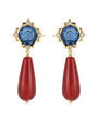 A pair of gold drop earrings featuring a faceted blue gemstone in a starburst setting with small clear accent stones, and a long teardrop-shaped red bead dangling below.