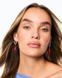 A close-up portrait of a woman with long brown hair wearing dangling earrings with red teardrop charms and silver-toned tops, against a plain white background.