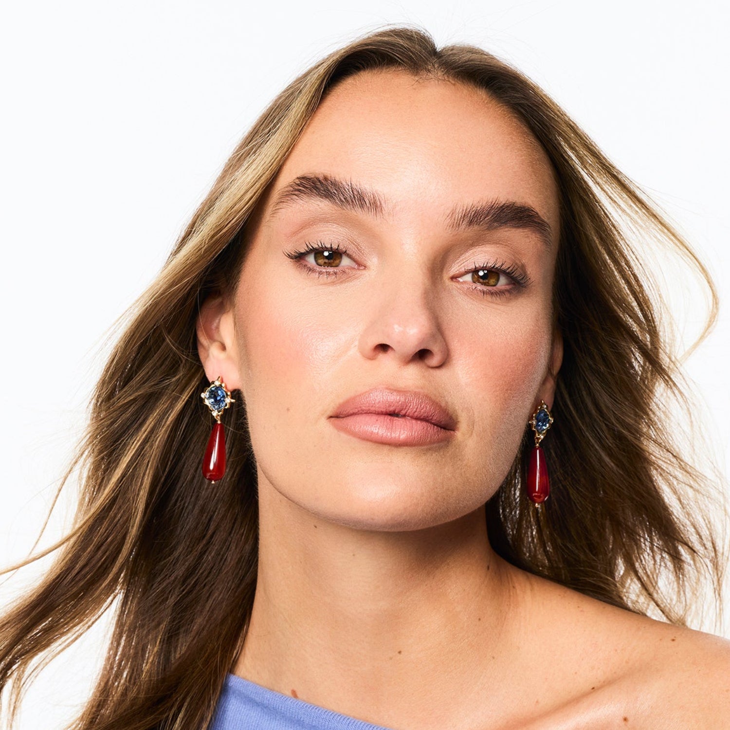 A close-up portrait of a woman with long brown hair wearing dangling earrings with red teardrop charms and silver-toned tops, against a plain white background.