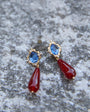 A pair of gold drop earrings featuring oval blue gemstones in scalloped settings and teardrop-shaped dark red dangling drops, displayed on a textured gray stone surface.