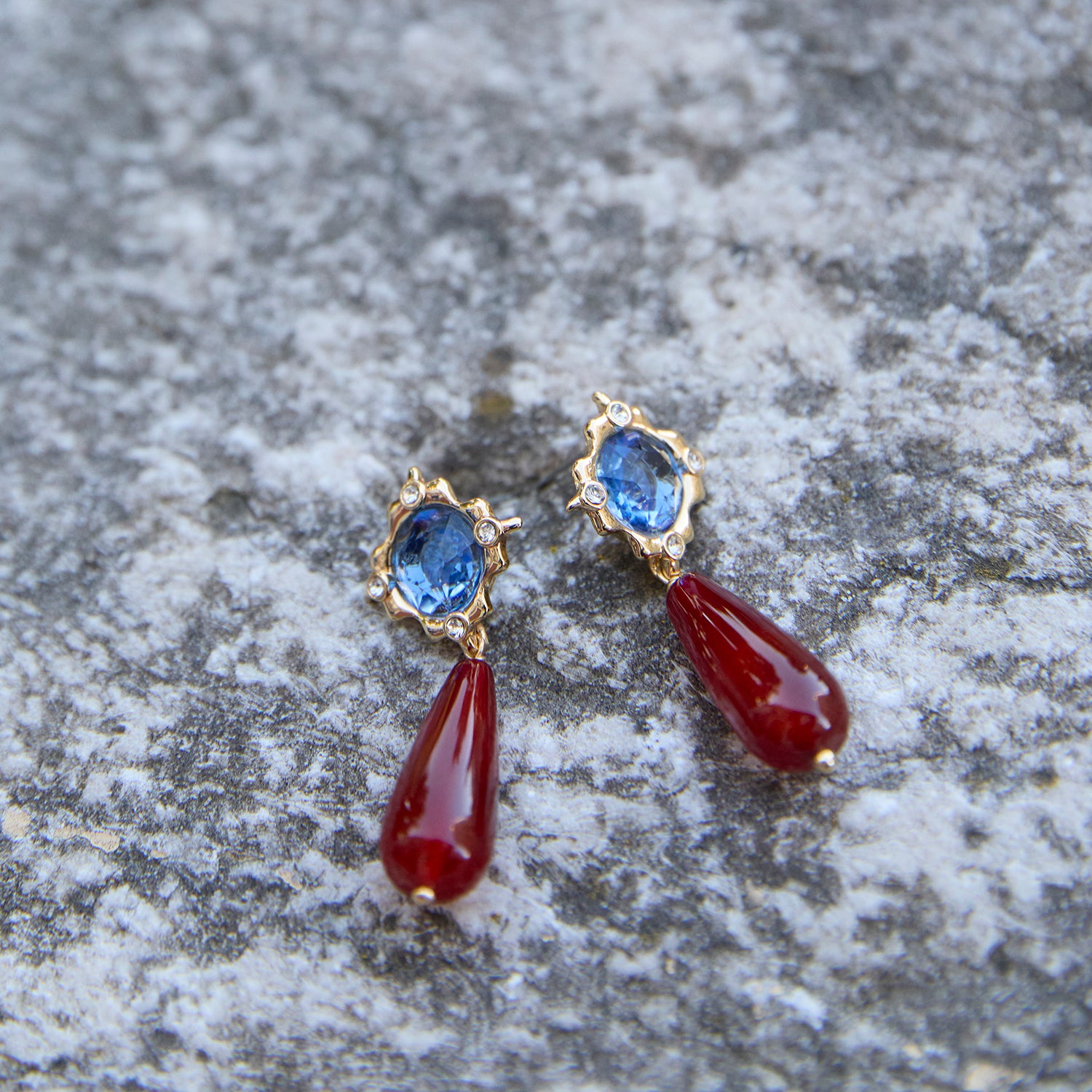 A pair of gold drop earrings featuring oval blue gemstones in scalloped settings and teardrop-shaped dark red dangling drops, displayed on a textured gray stone surface.