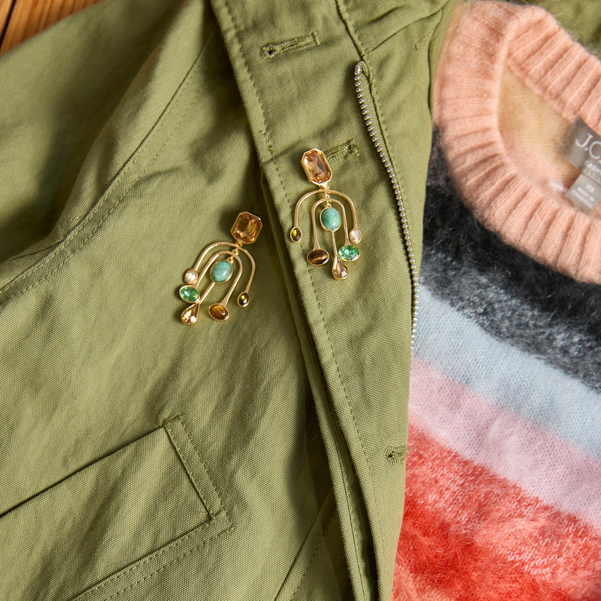 A close-up flat lay of a pair of gold-toned arch-shaped earrings with turquoise and amber stones, styled on an olive green jacket over a striped knit sweater.