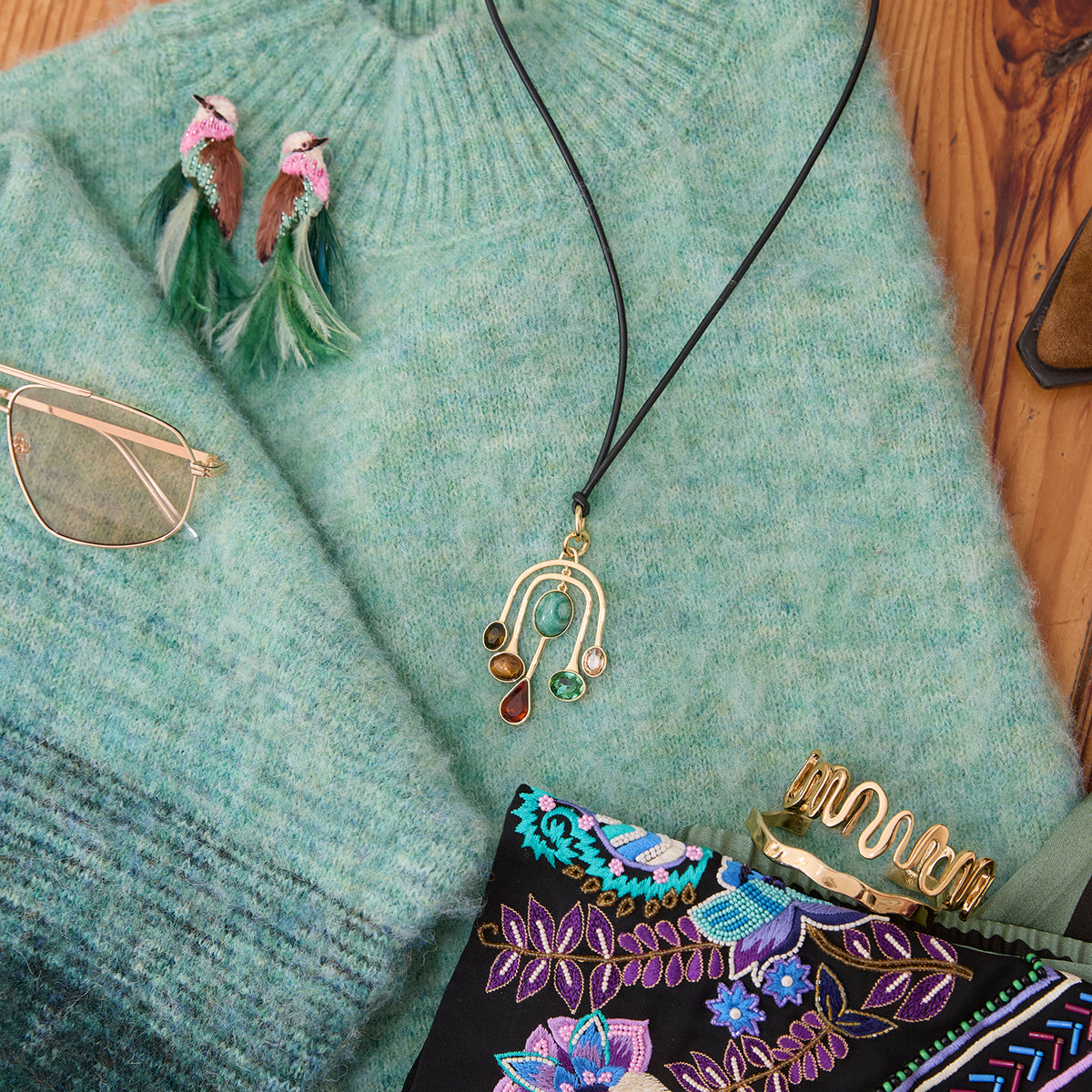 A gold crescent moon pendant necklace with green and brown beads on a black cord, styled on a light blue-green knit sweater alongside sunglasses, a feathered earring, and a patterned clutch on a wooden surface.