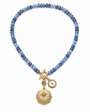 A beaded necklace featuring light and dark blue stones with gold-toned charms, including a compass and a small white stone charm, displayed on a clean white background.