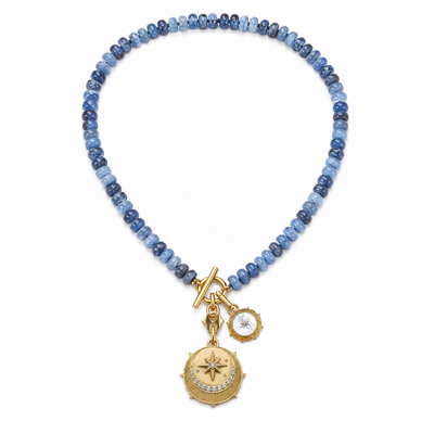 A beaded necklace featuring light and dark blue stones with gold-toned charms, including a compass and a small white stone charm, displayed on a clean white background.