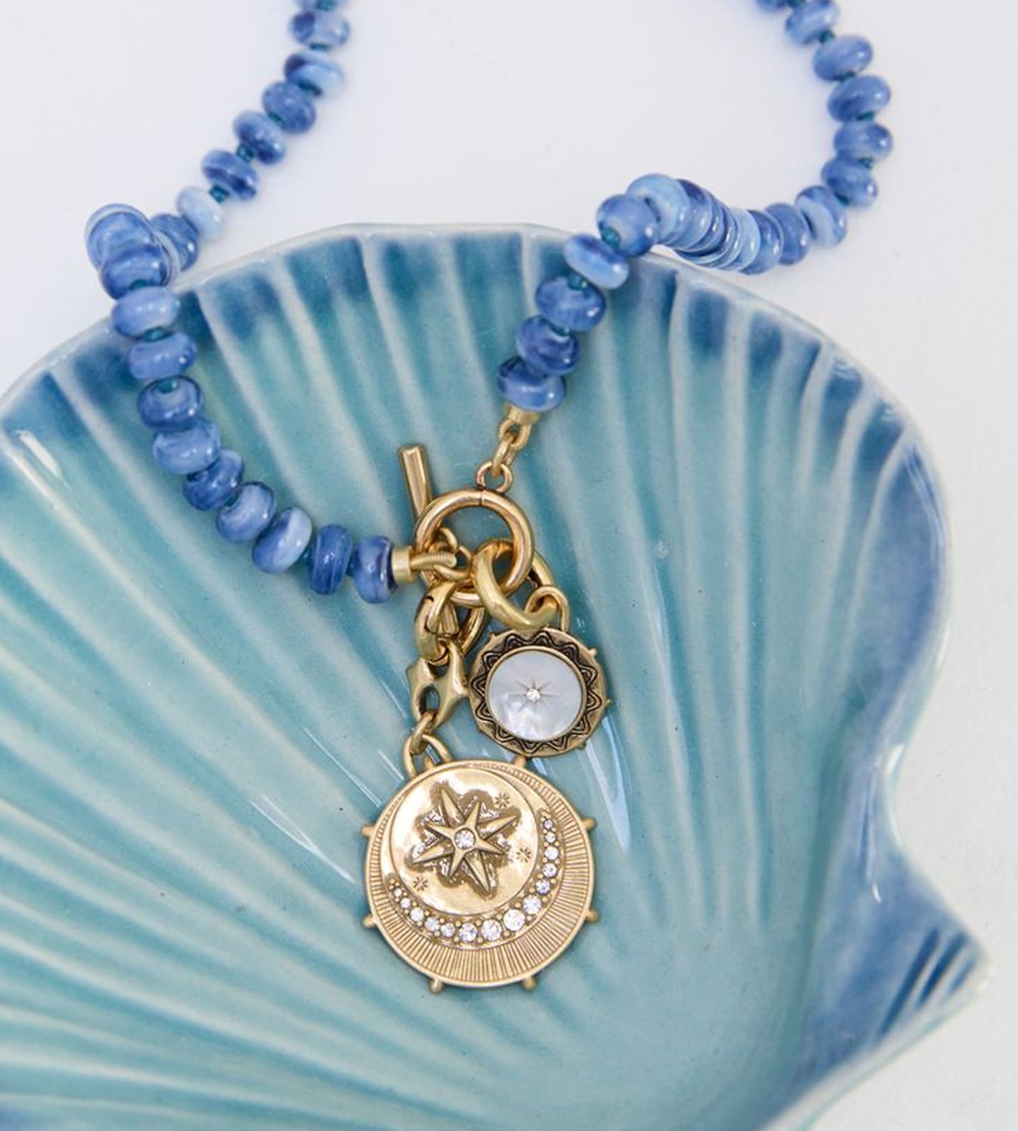 A blue beaded necklace with gold-toned charms, including a moon and star pendant, displayed on a light blue ceramic shell dish against a white background.