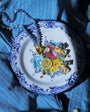 A close-up of jewelry displayed on a decorative white plate with blue floral border and painted flowers, placed on top of a blue striped button-up shirt. The jewelry includes a beaded necklace, a gold-toned pendant, and several small earrings with blue, yellow, and metallic details.