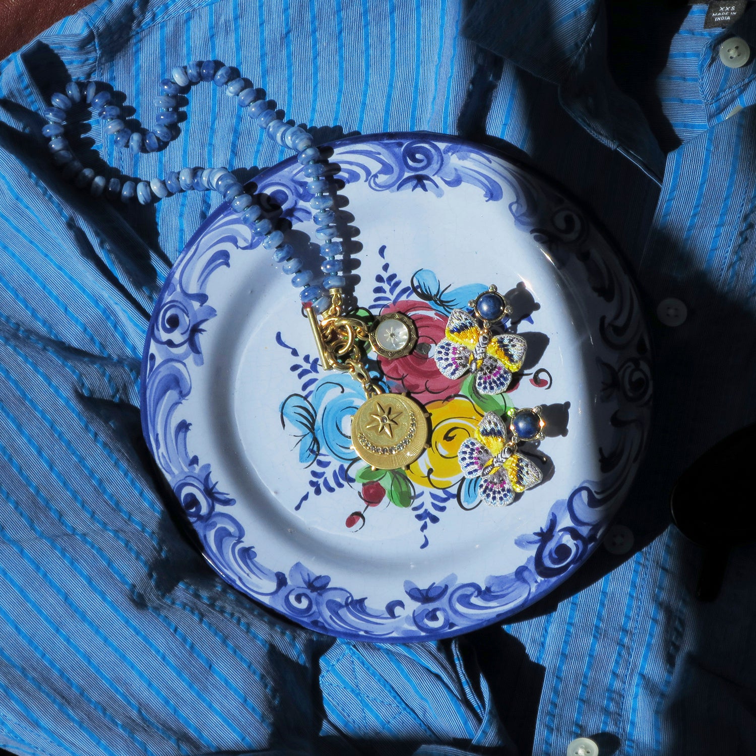 A close-up of jewelry displayed on a decorative white plate with blue floral border and painted flowers, placed on top of a blue striped button-up shirt. The jewelry includes a beaded necklace, a gold-toned pendant, and several small earrings with blue, yellow, and metallic details.