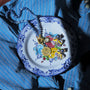 A close-up of jewelry displayed on a decorative white plate with blue floral border and painted flowers, placed on top of a blue striped button-up shirt. The jewelry includes a beaded necklace, a gold-toned pendant, and several small earrings with blue, yellow, and metallic details.