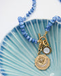 A beaded necklace with light blue round beads and a gold-toned pendant featuring a crescent moon and star design, displayed on a light blue scalloped dish against a white background.
