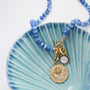 A beaded necklace with light blue round beads and a gold-toned pendant featuring a crescent moon and star design, displayed on a light blue scalloped dish against a white background.