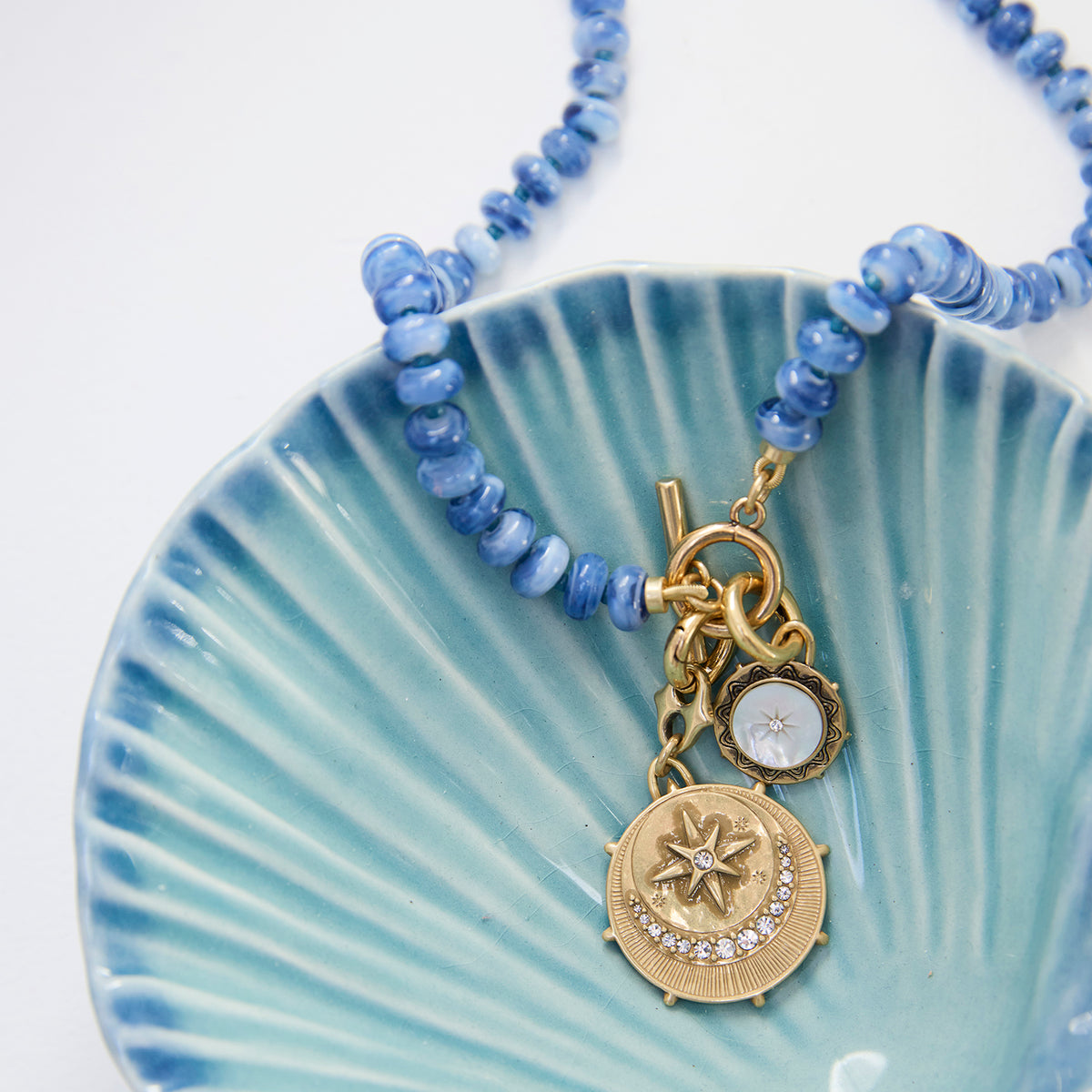 A beaded necklace with light blue round beads and a gold-toned pendant featuring a crescent moon and star design, displayed on a light blue scalloped dish against a white background.
