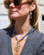 A woman wearing a blue beaded necklace with gold-toned pendants and matching drop earrings, photographed outdoors in bright sunlight.