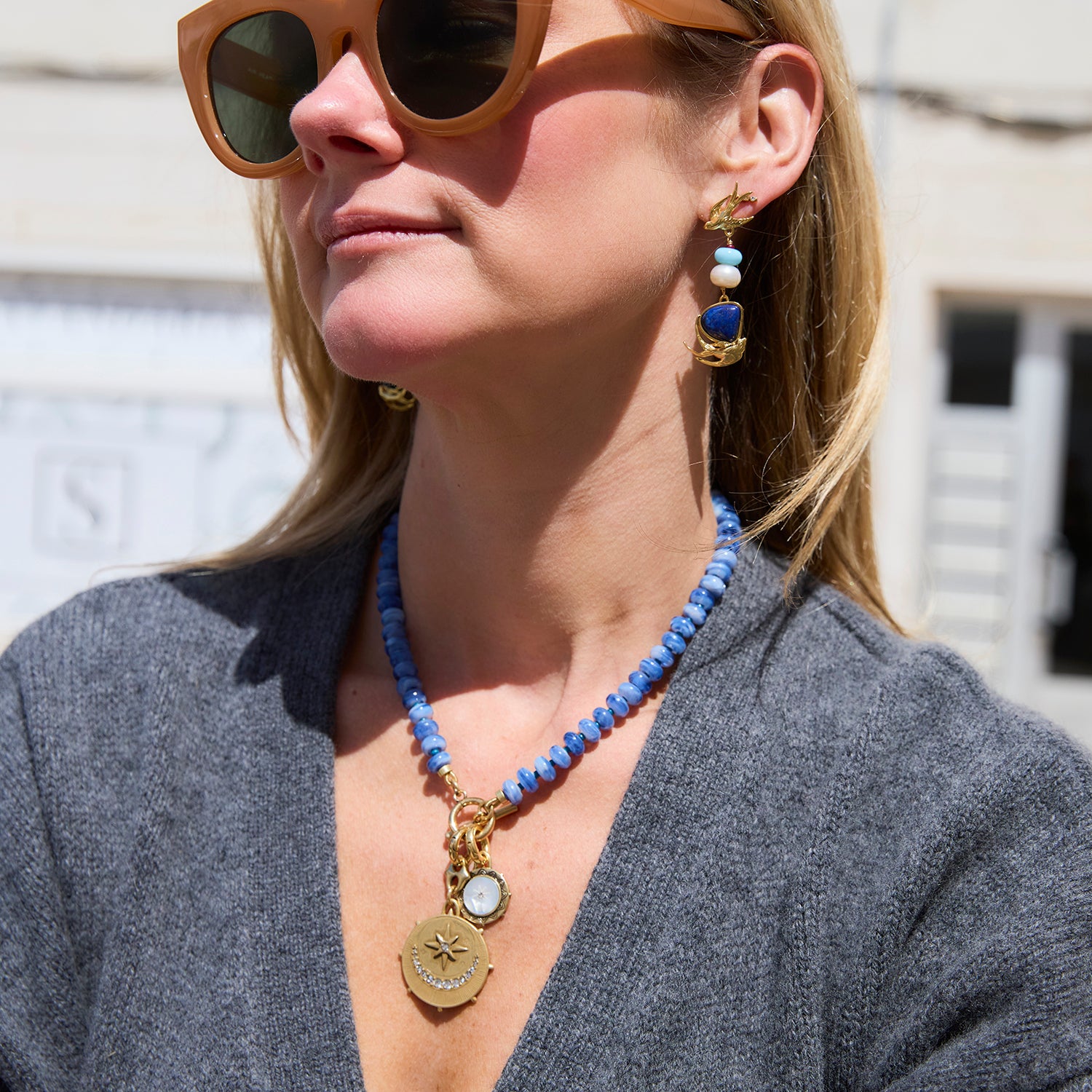 A woman wearing a blue beaded necklace with gold-toned pendants and matching drop earrings, photographed outdoors in bright sunlight.