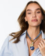 A woman wearing a layered necklace with a blue beaded top, gold-toned chain, and a large oval blue pendant with gold detailing, paired with a light blue button-down shirt against a white background.