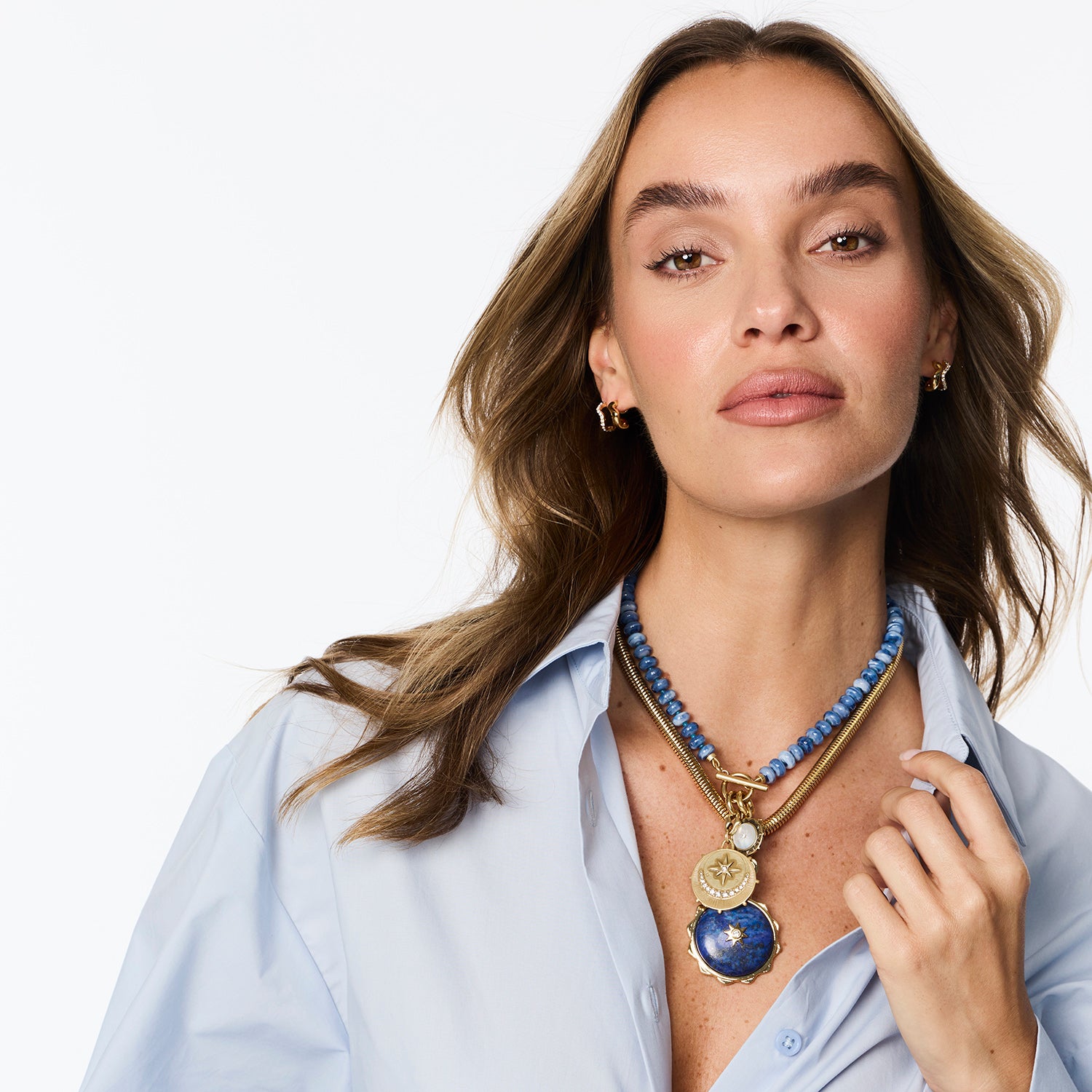 A woman wearing a layered necklace with a blue beaded top, gold-toned chain, and a large oval blue pendant with gold detailing, paired with a light blue button-down shirt against a white background.