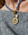 A close-up of a person wearing a beaded necklace with a gold-toned compass pendant and a small round white stone charm, styled over a gray knit top.