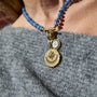 A close-up of a person wearing a beaded necklace with a gold-toned compass pendant and a small round white stone charm, styled over a gray knit top.
